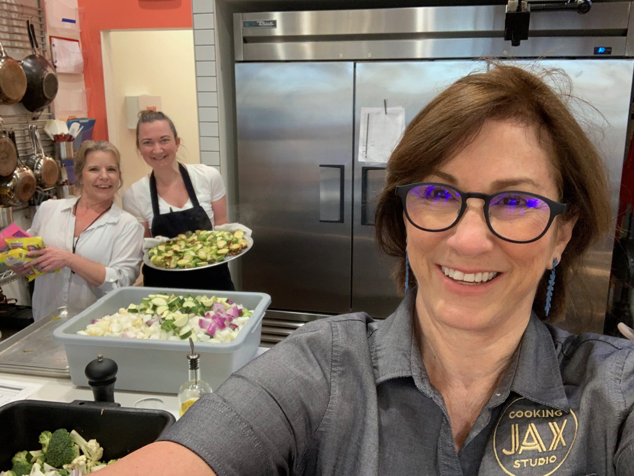 A selfie from the angle of Terri, owner of JAX Cooking Studio. Two female employees prepare food in the background.