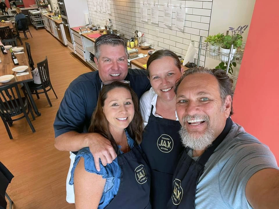 A selfie of four JAX Cooking Studio customers, wearing aprons in the studio. Kitchen environment in the background.
